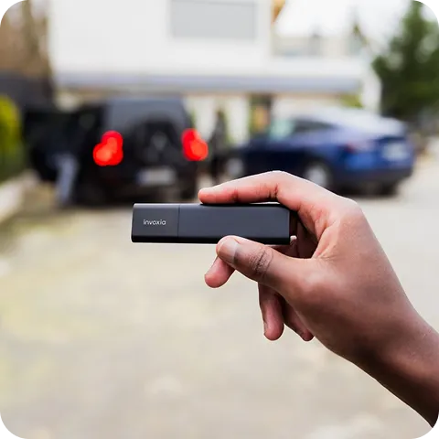 A Classic GPS Tracker held in a hand, with a blurred background showing two cars in a courtyard