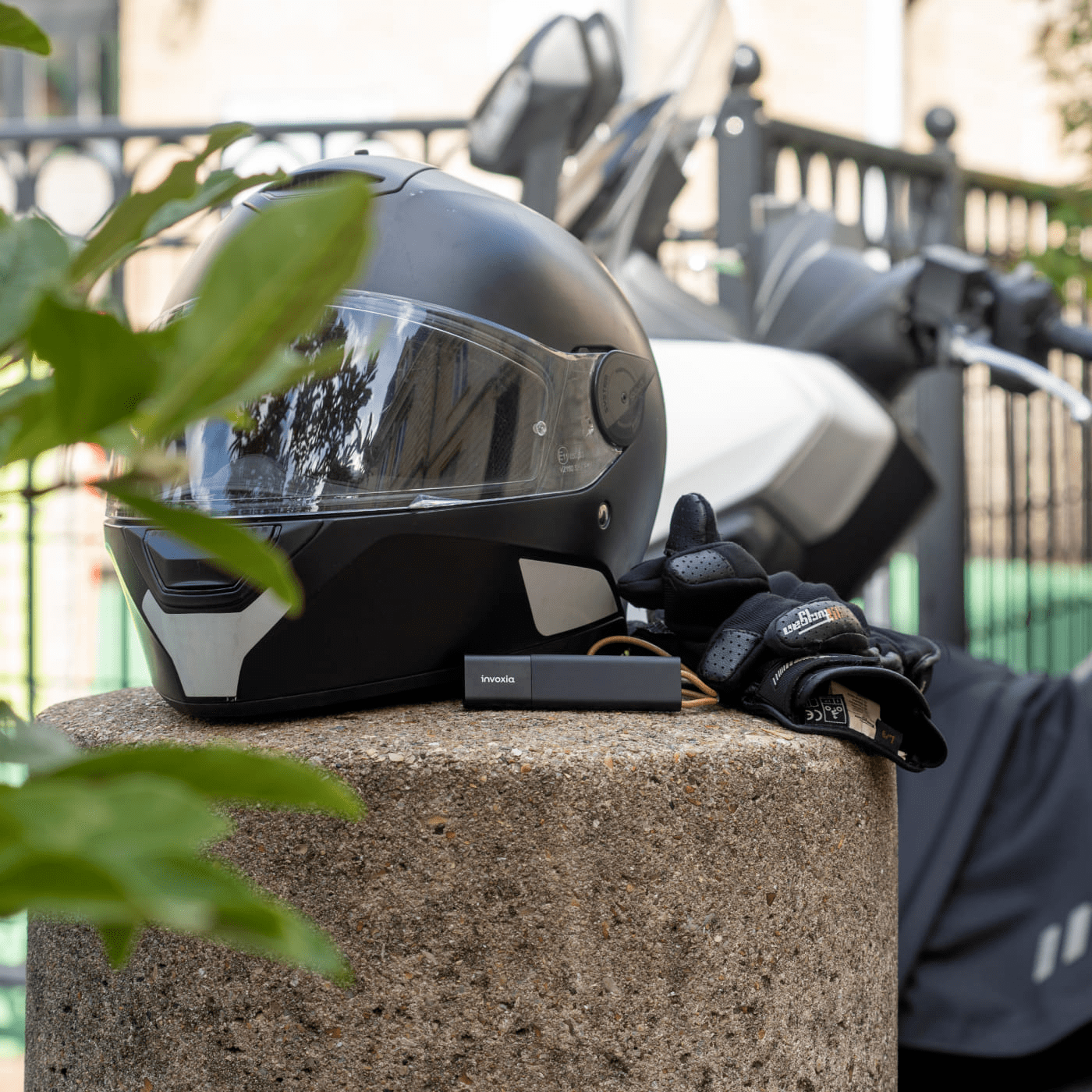 Invoxia GPS-Tracker neben einem Motorradhelm, im Hintergrund ein Motorroller